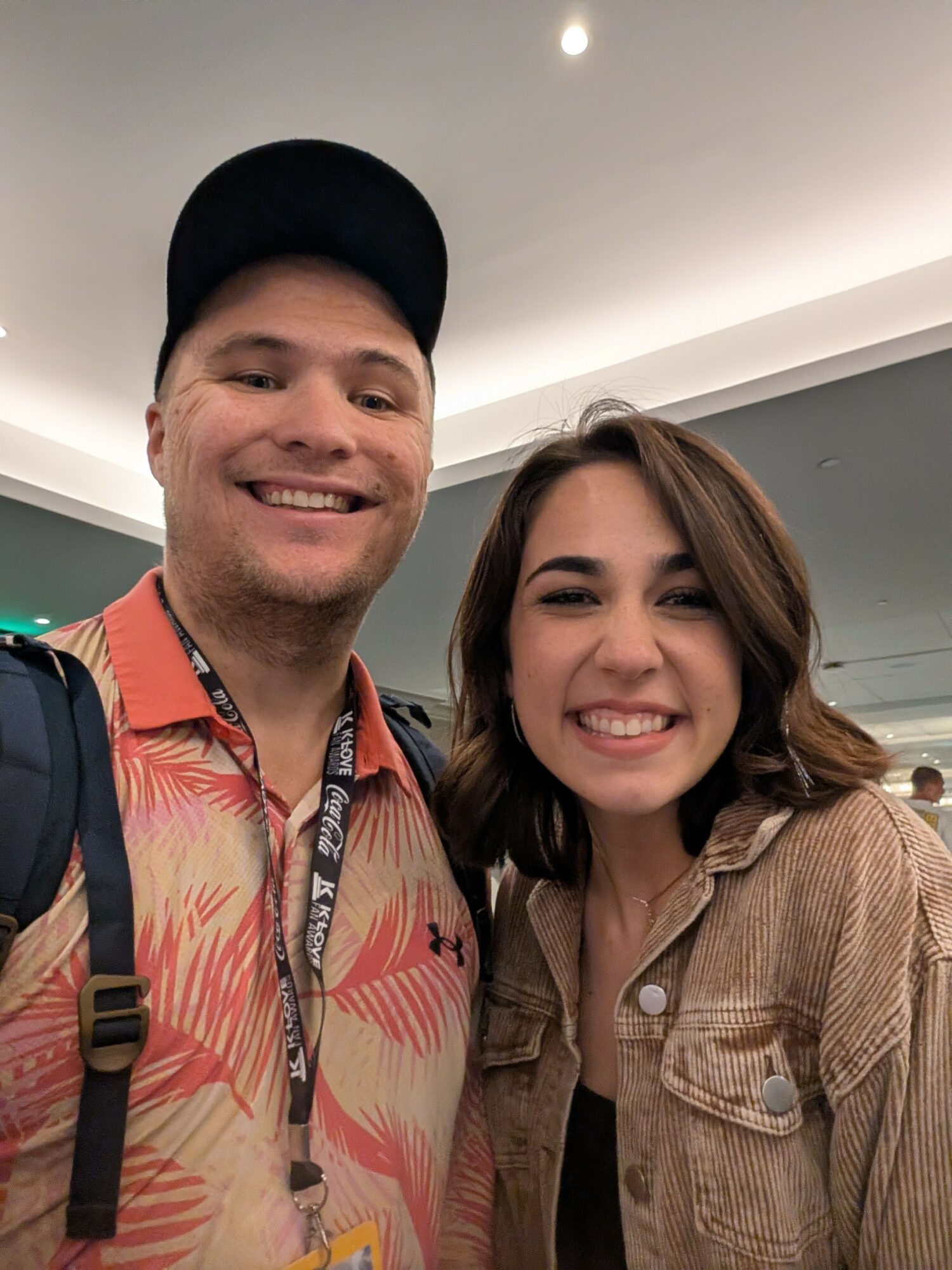 Two smiling people, a man and a woman, standing close together indoors, both facing the camera.