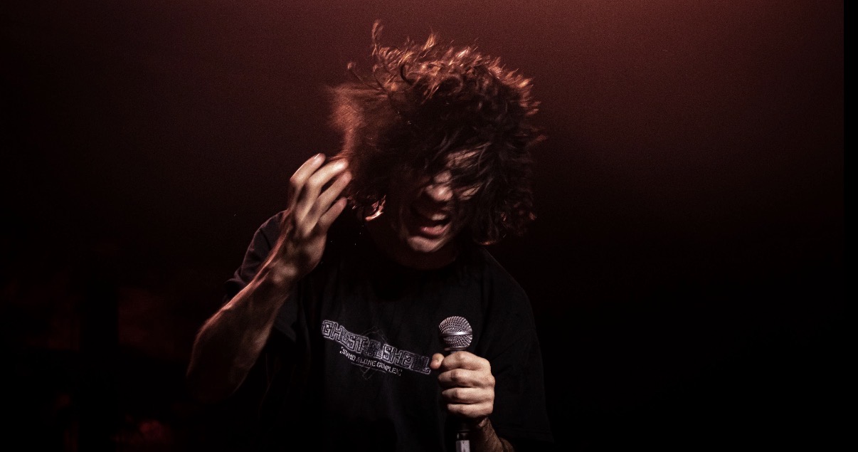 Person with curly hair holding a microphone, head slightly tilted, dark background, red lighting, expressive pose.