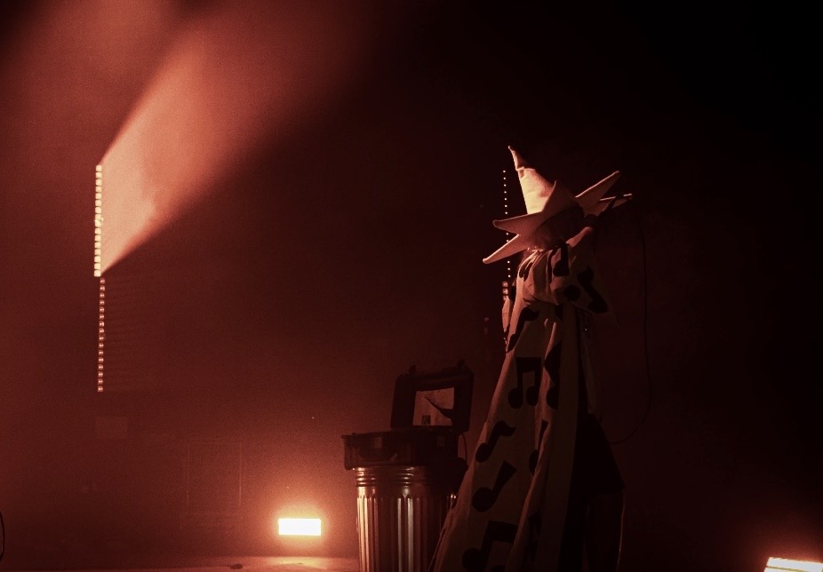 Stage with a person in costume, illuminated by warm lighting, and a large screen in the background.