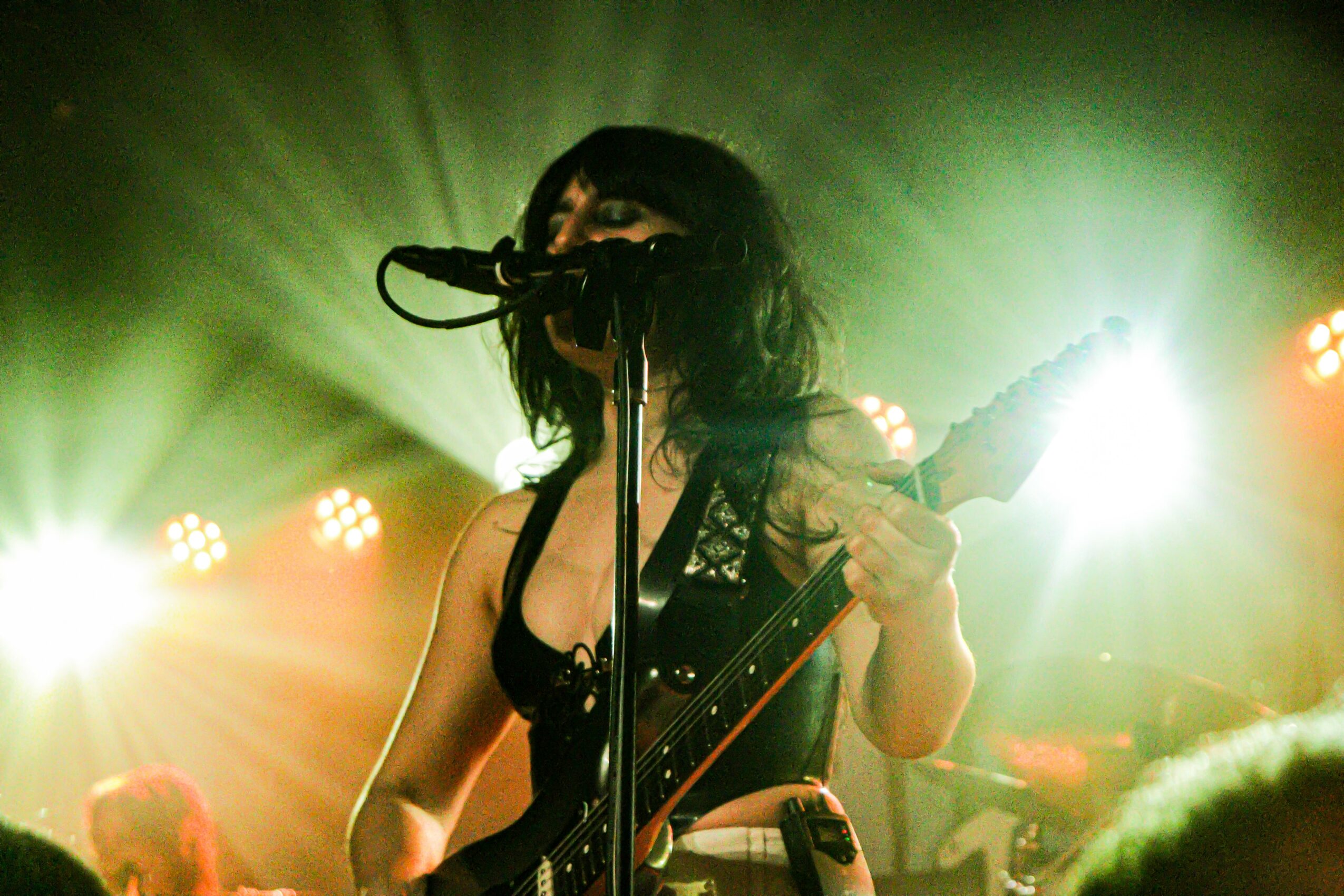 Female musician singing into microphone with guitar on stage, bright lights behind her, dark background.