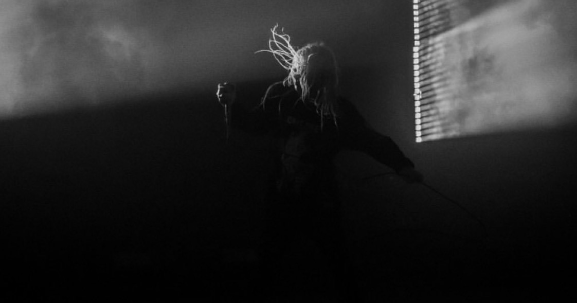 Silhouette of a person with long hair holding a stick or cane, standing in a dark room with light coming through blinds.