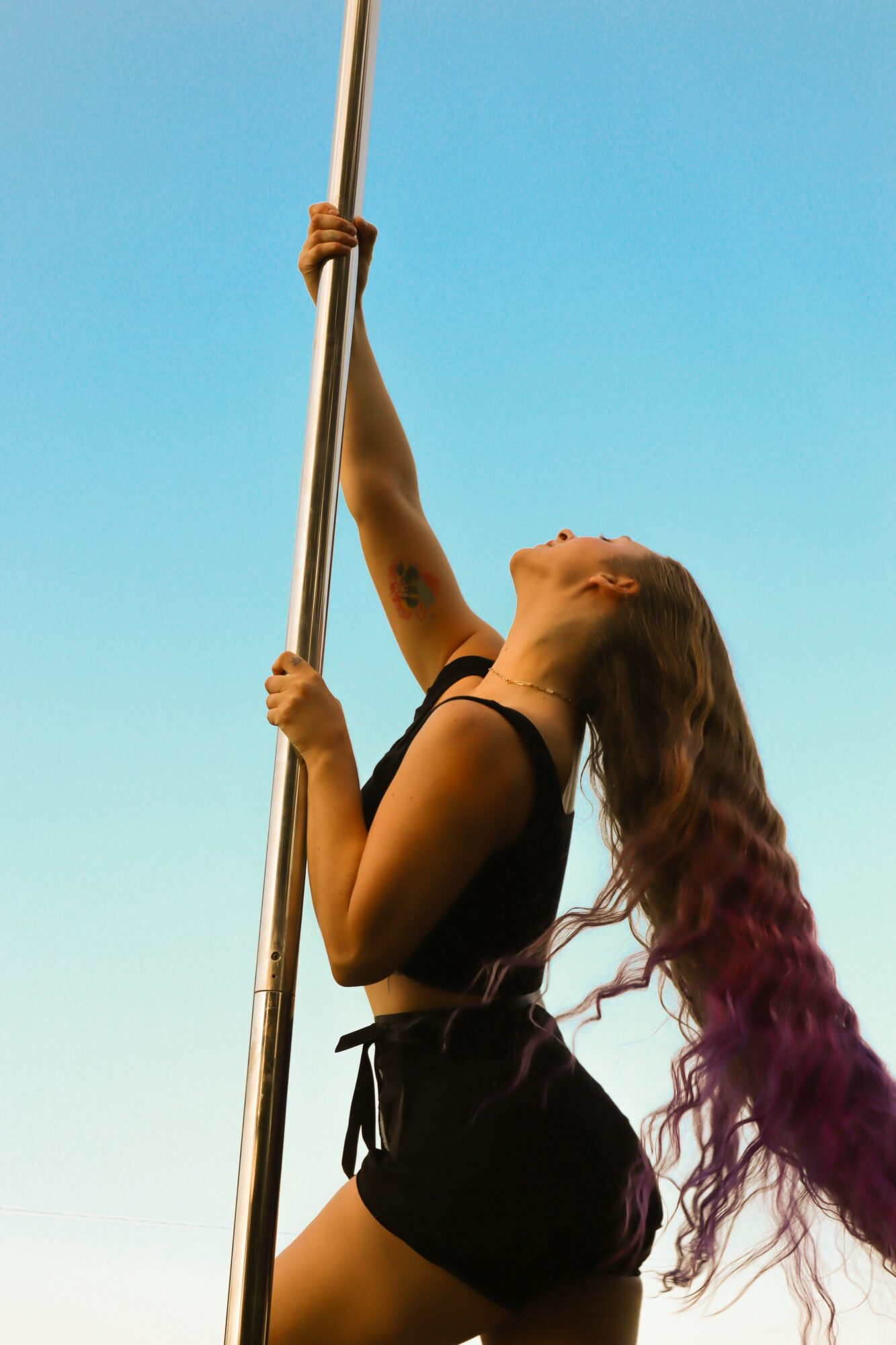 Woman with long, wavy hair holding a pole against a clear blue sky, looking upward, wearing a black outfit.