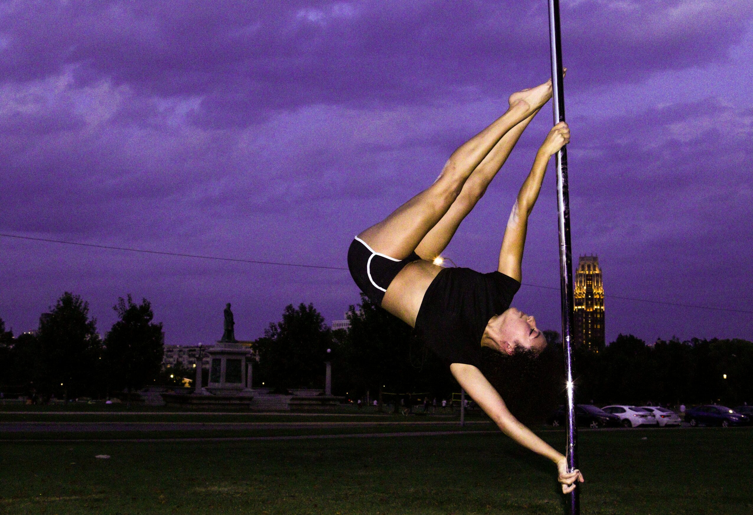 Person performing pole dance move outdoors at dusk with purple sky, trees, and cityscape in background.