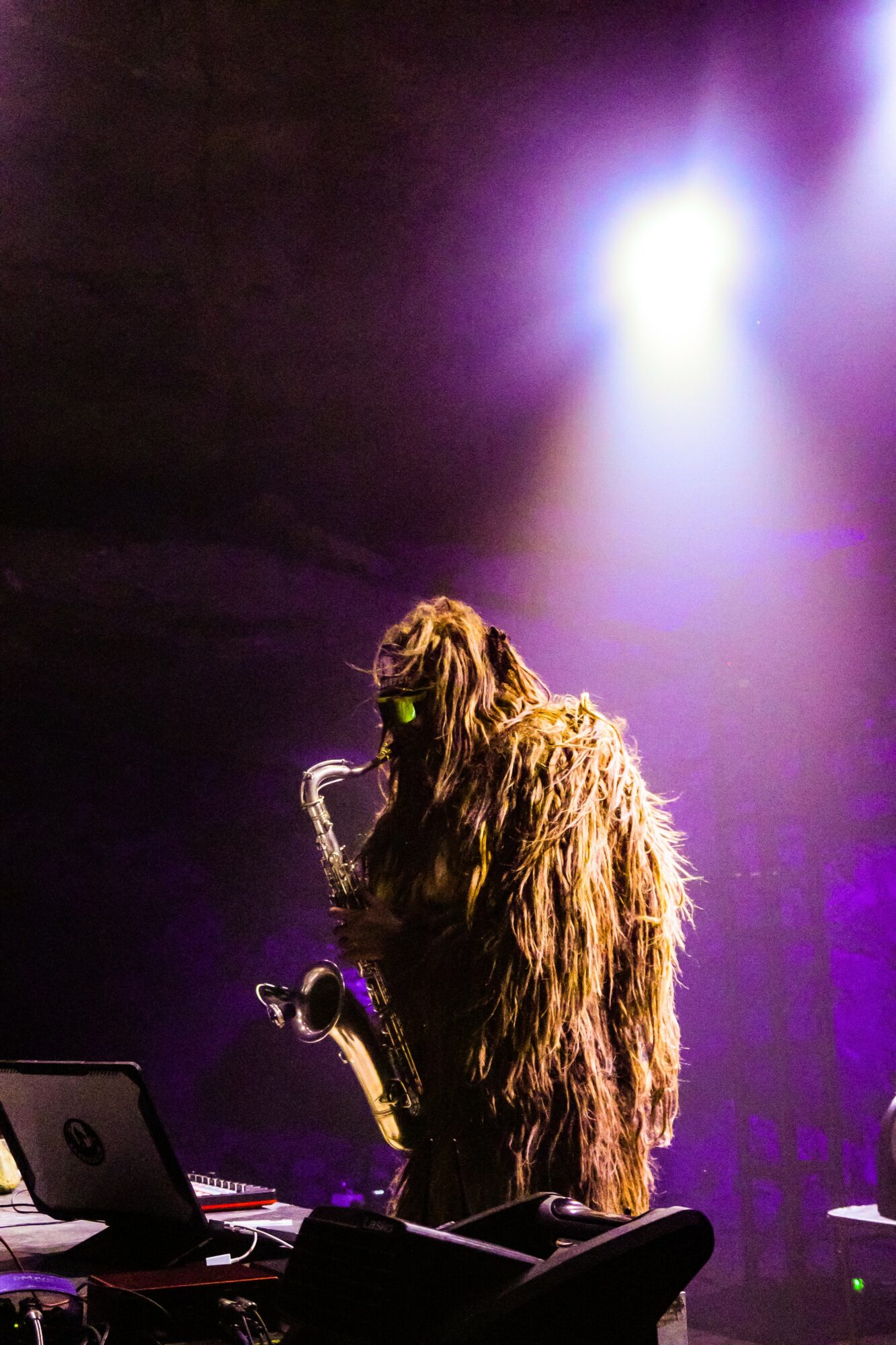 Person dressed as a furry creature playing a saxophone on stage with bright purple and white lights.