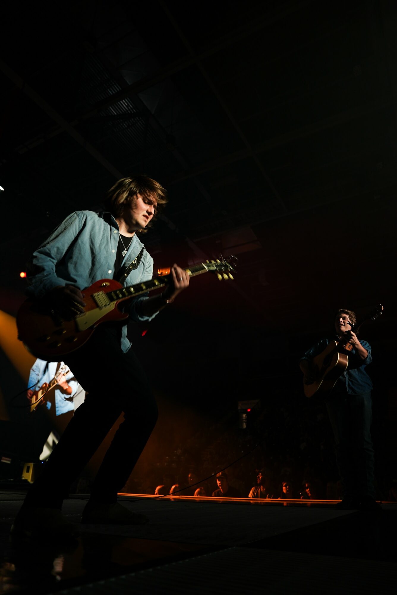 Musician playing guitar on stage with dark background and stage lighting, audience visible at bottom.