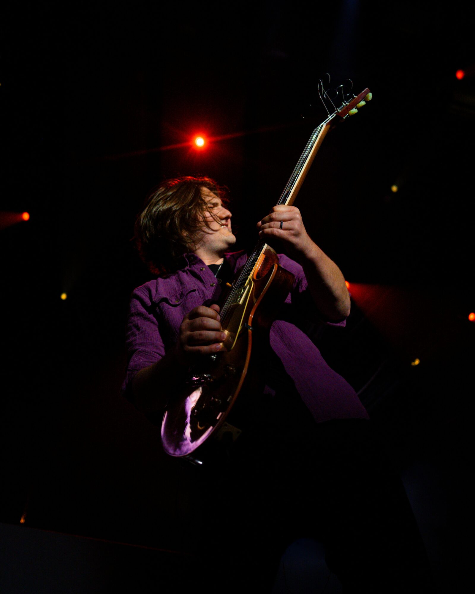 Person playing an electric guitar on stage with red and yellow lights in the background, wearing a purple jacket.