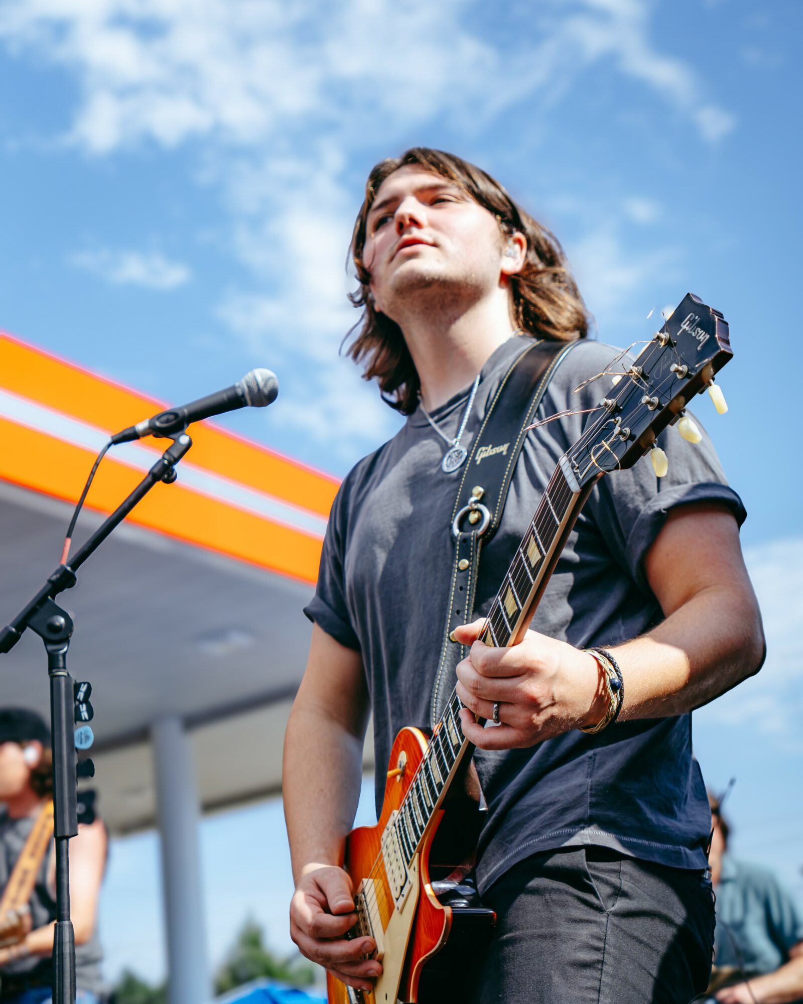 Young man with long hair playing an electric guitar outdoors under a blue sky with clouds, standing near a microphone.