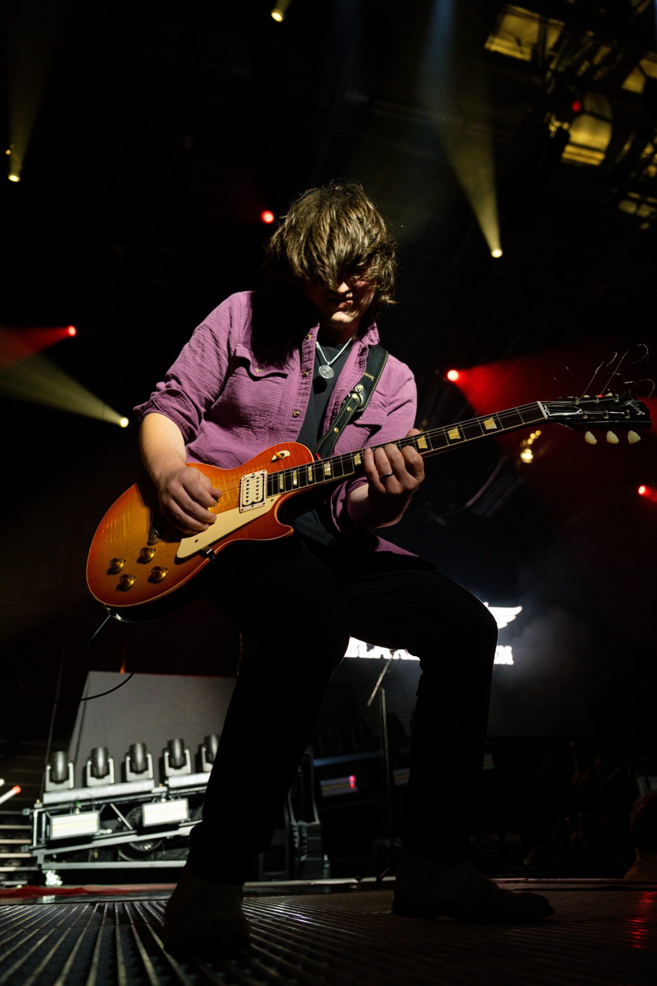 Person playing electric guitar on stage with colorful lighting, wearing a purple shirt and dark pants.
