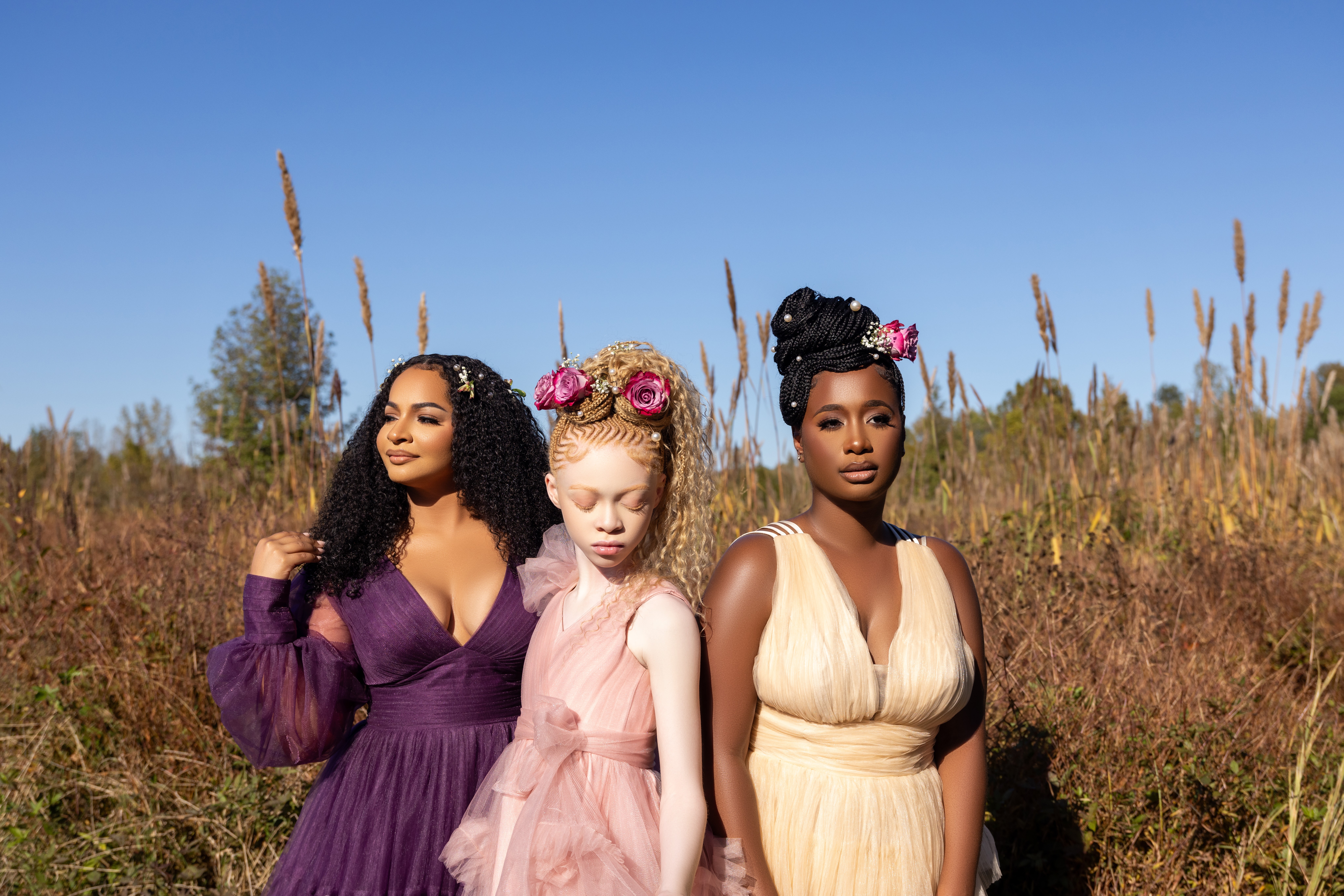 Three women standing outdoors in a field with tall grass and blue sky, wearing colorful dresses and floral headbands.