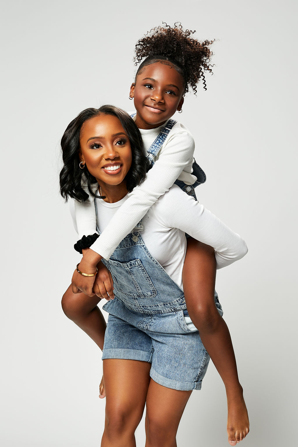 Young girl piggybacking on woman, both smiling, against a plain white background.