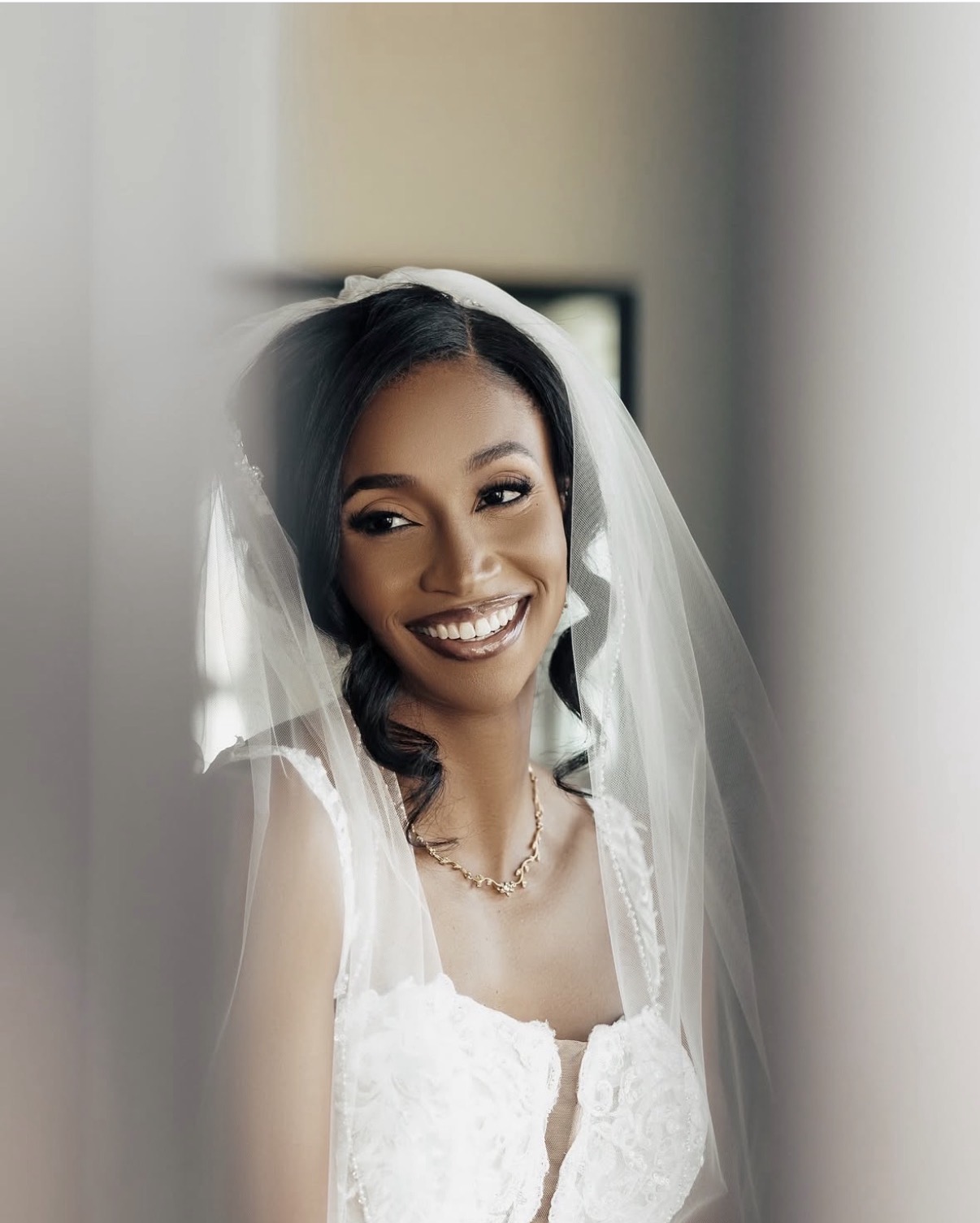 Smiling woman in wedding dress and veil, looking out a window, with soft lighting and blurred background.