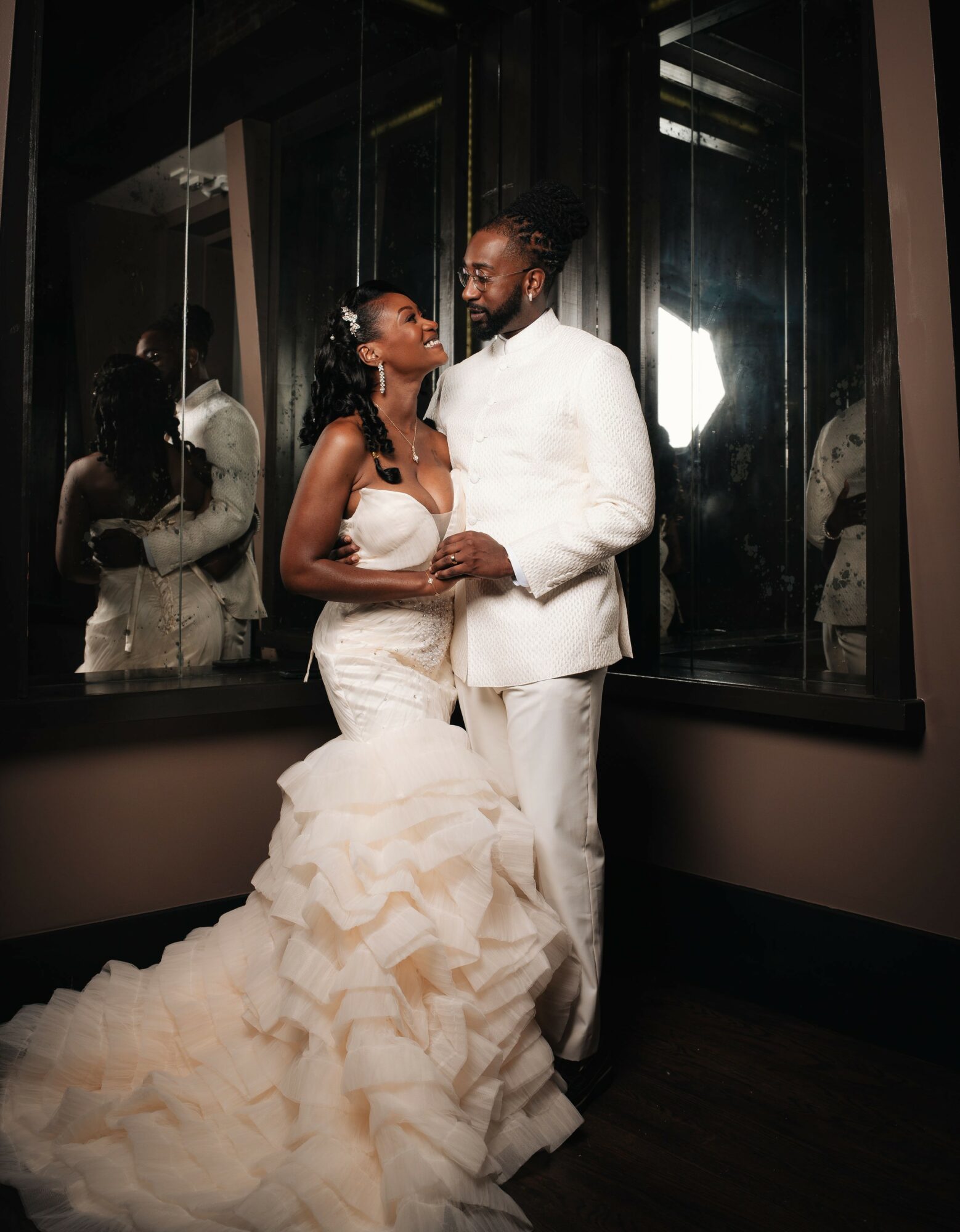 A couple in wedding attire stands close together, smiling, in front of large windows with reflections. The bride wears a ruffled gown, the groom a white suit.