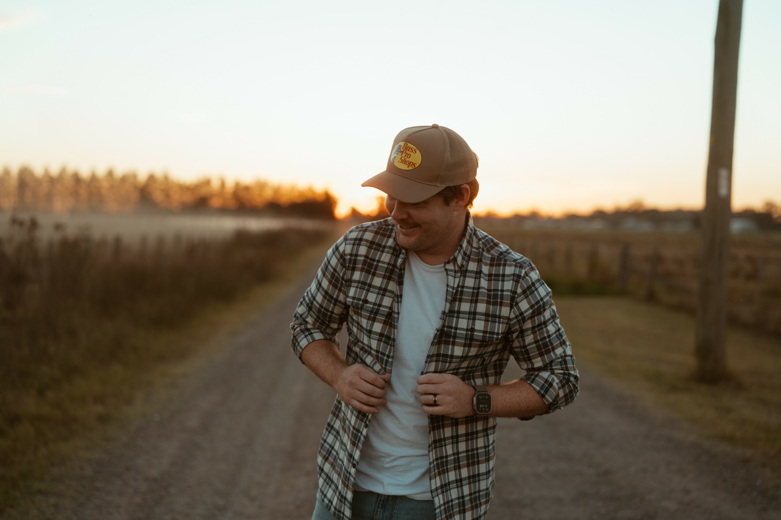 Person wearing a plaid shirt and cap walking on a dirt path during sunset, holding a backpack strap, smiling.