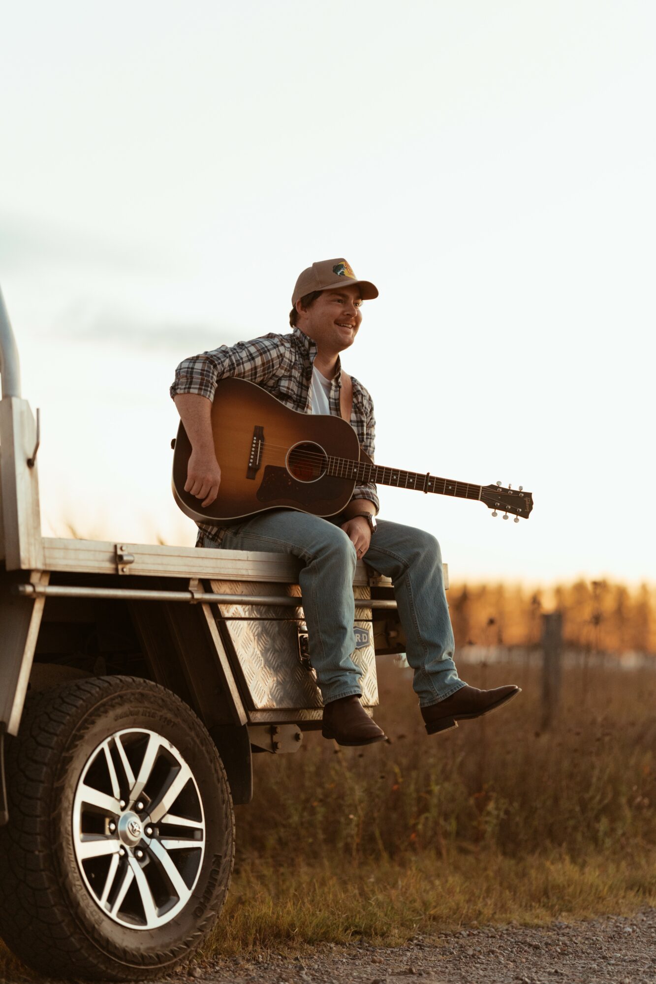 Person sitting on vehicle with a guitar, outdoors during sunset, wearing a hat and plaid shirt.