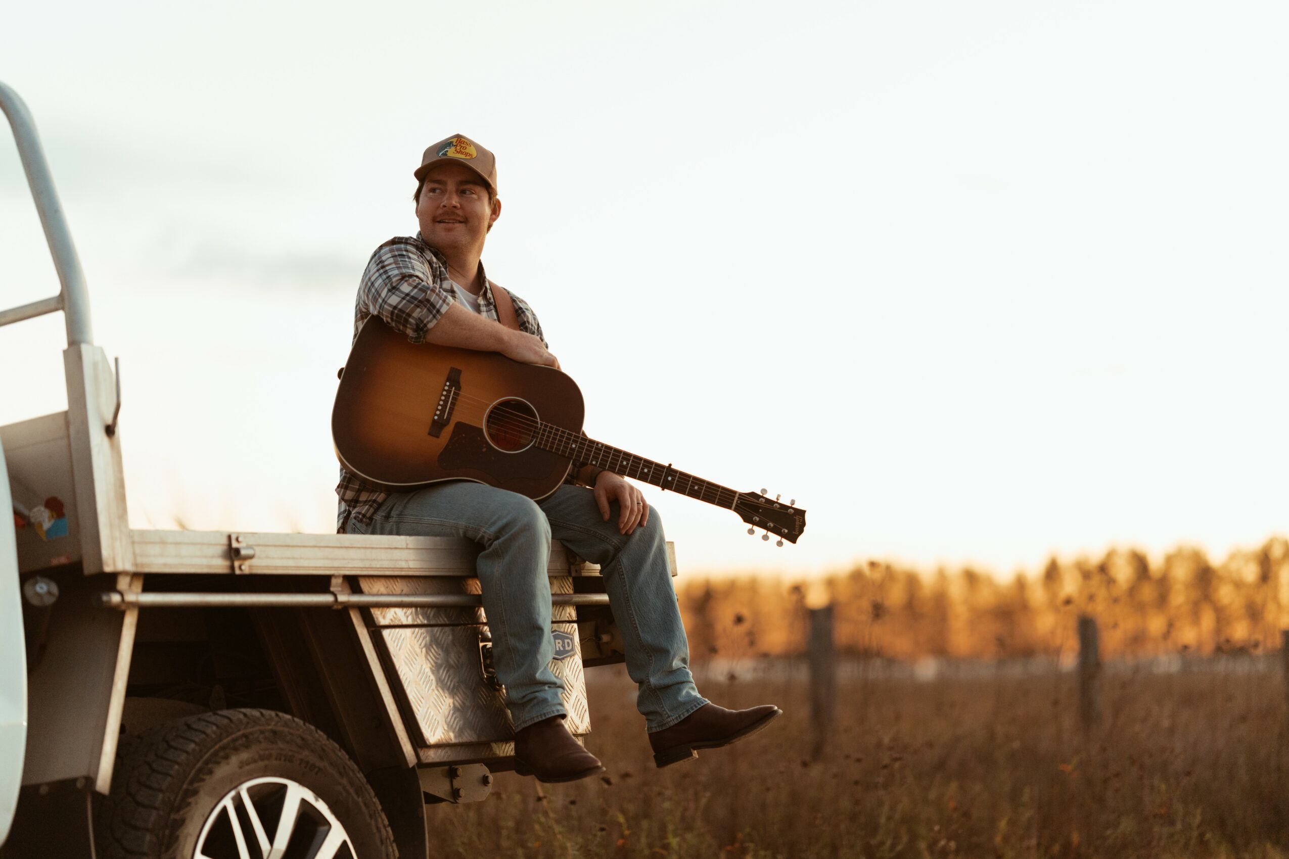 Man sitting on truck bed holding a guitar outdoors, with a field and sky in the background.
