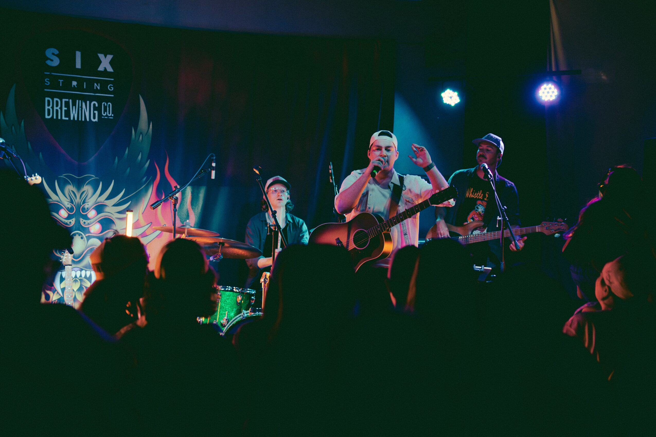 Band performs on stage with three musicians, one singing and playing guitar, audience silhouettes in foreground, colorful stage lights, and a backdrop with text.