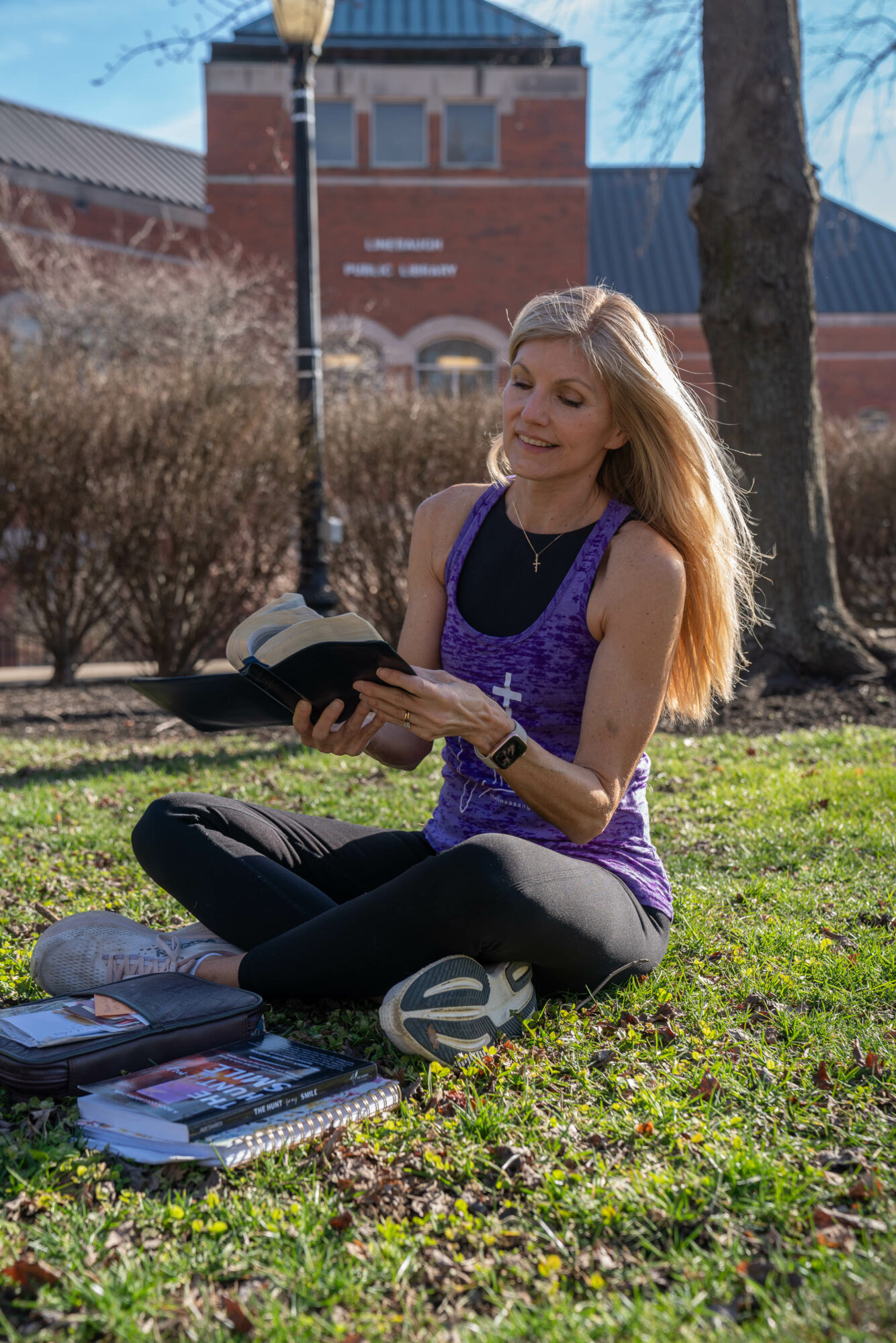 Woman sitting on grass reading a book outdoors, with books and a backpack nearby, trees and a building in background.