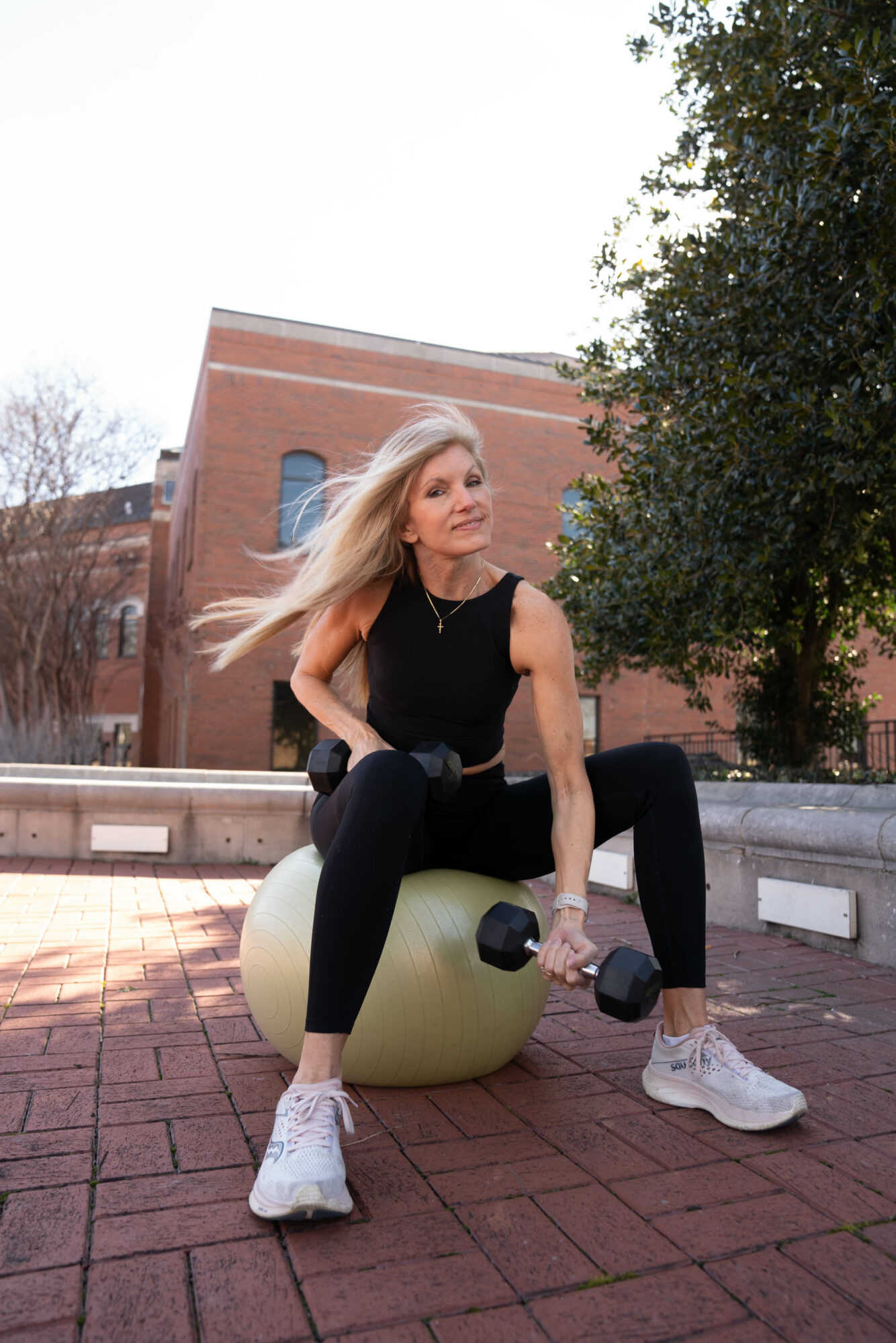 Woman with long blonde hair sitting on a yellow exercise ball outdoors, holding a dumbbell, with trees and brick building in background.