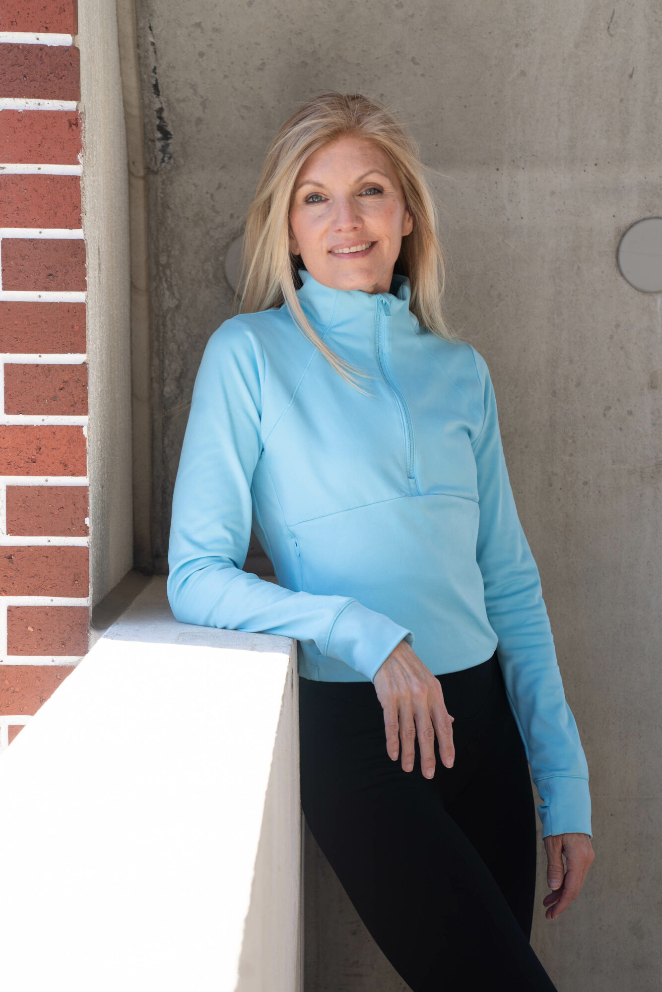 Woman with blonde hair in a blue jacket leaning against a wall, smiling outdoors.