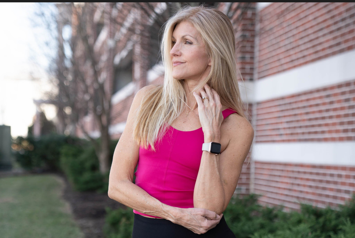Woman with long blonde hair wearing a pink sleeveless top outdoors near a brick building, looking to the side, touching her neck.
