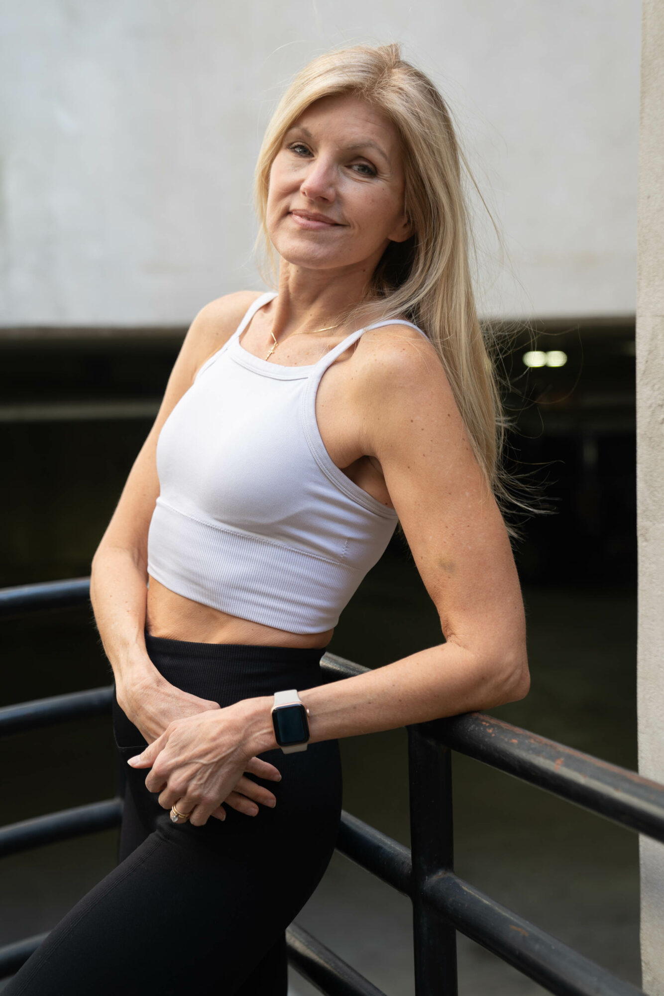 Woman with long blonde hair in a white crop top and black pants leaning on a railing outdoors.