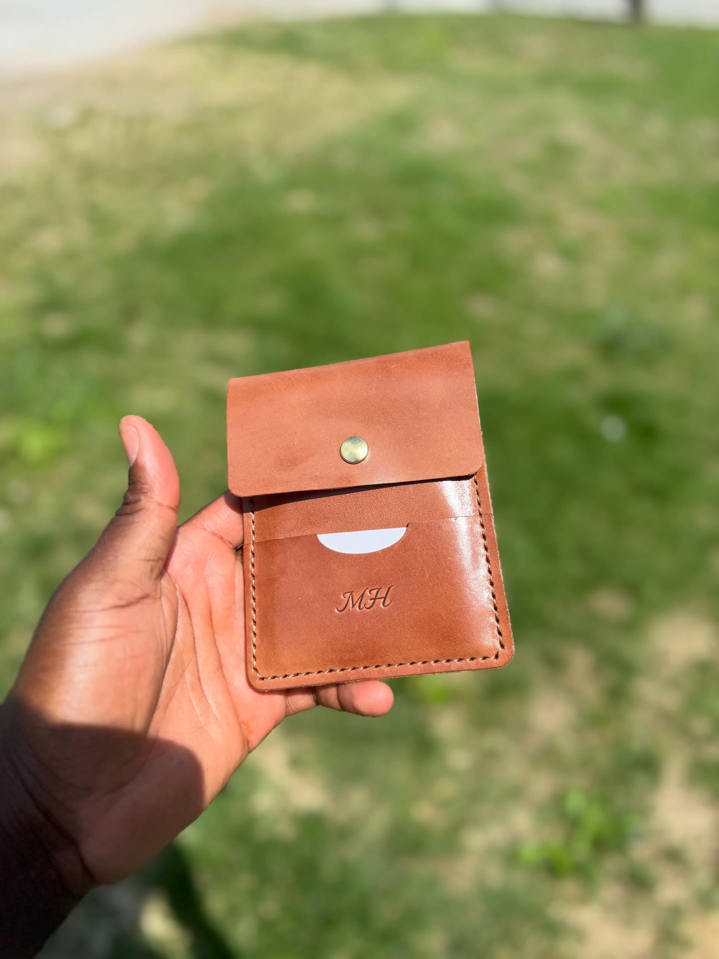 Hand holding a brown leather wallet with a flap and snap button, outdoors on grass.