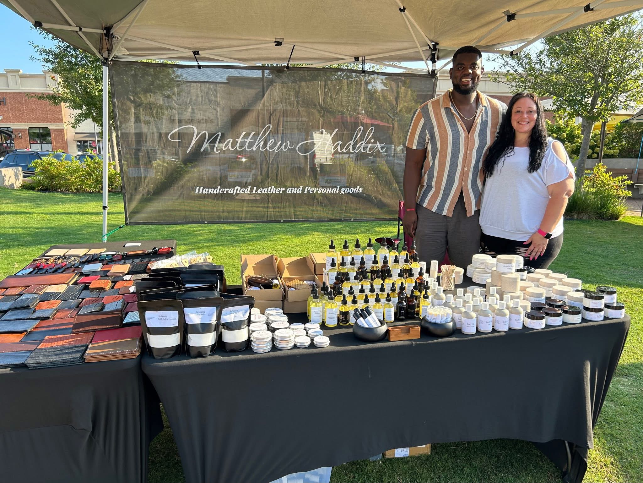 Two people standing behind a table with skincare products at an outdoor market, with a banner in the background.