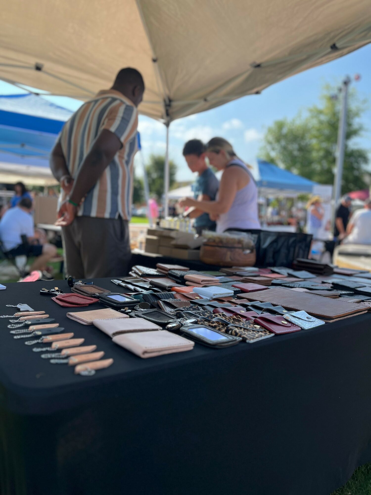 People browsing a table with various jewelry and accessories at an outdoor market under a tent, with other tents and trees in background.
