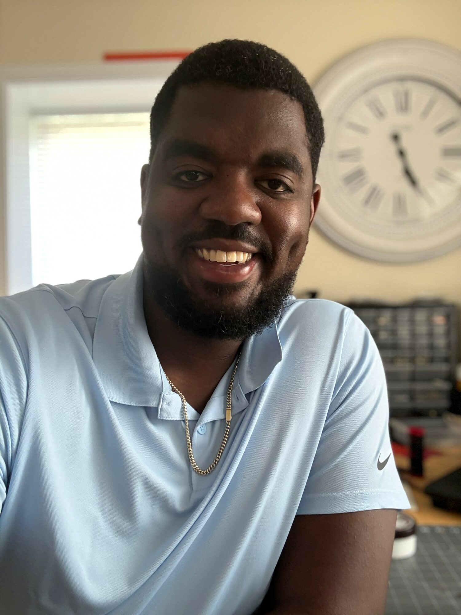 Smiling man with short hair and beard wearing a light blue shirt indoors, with a clock and window in the background.