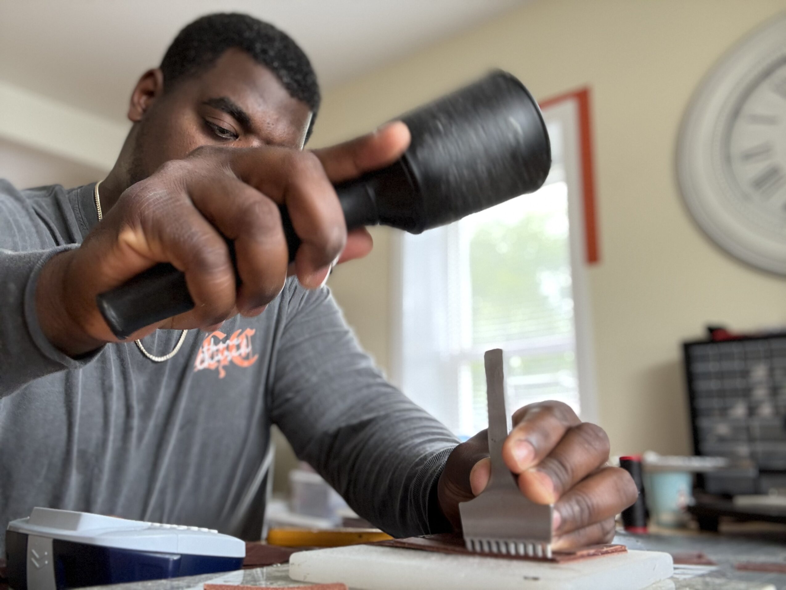 Person using a hammer on a small object on a table, with a clock and window in the background.