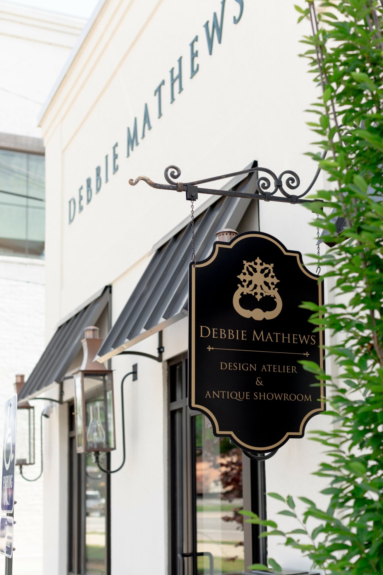 Storefront with sign for Debbie Mathews Design Atelier and Antique Showroom, awning, and greenery, viewed from below.
