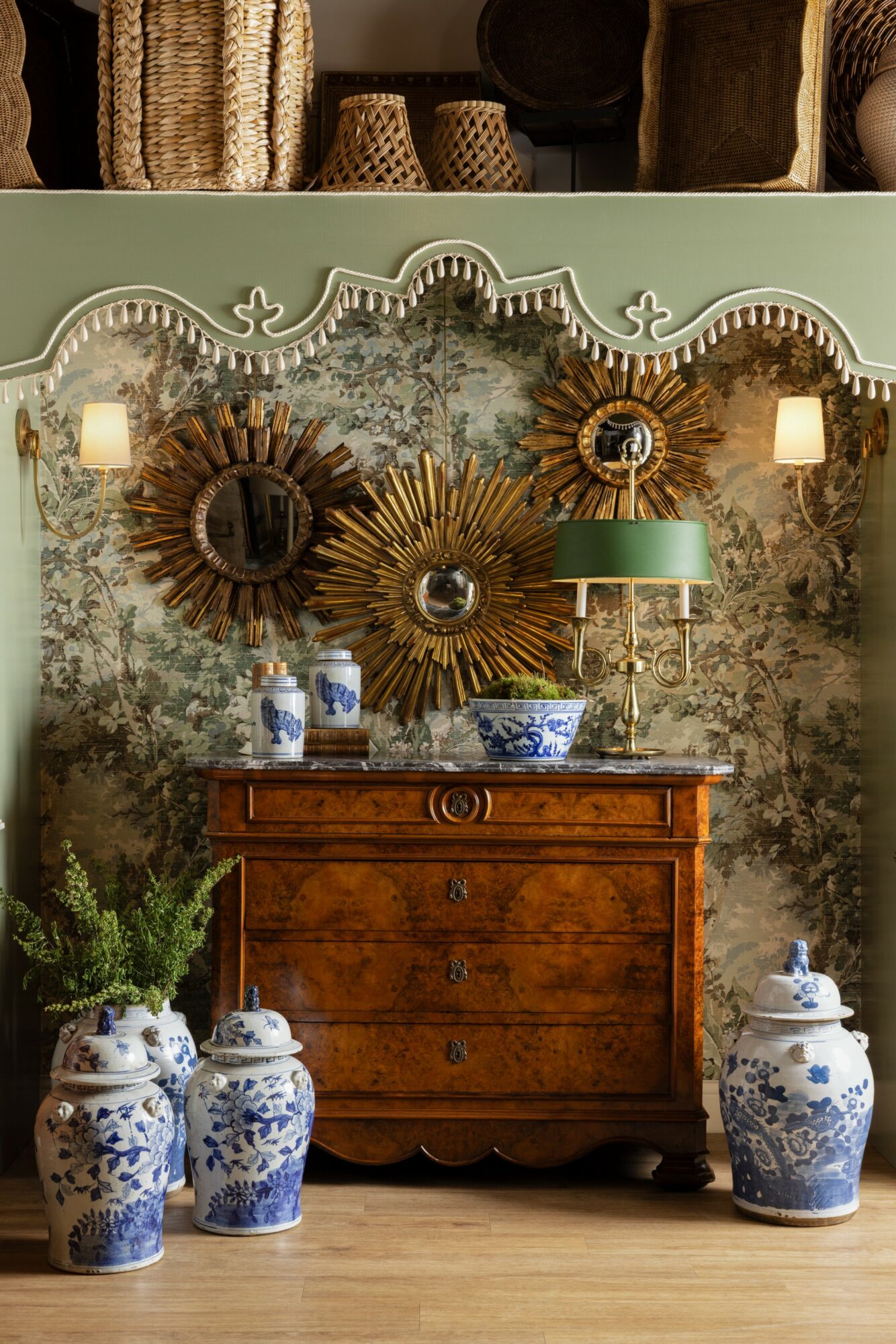 Decorative wall with three sunburst mirrors, a green lamp, and a wooden chest, flanked by blue and white vases, in a room.