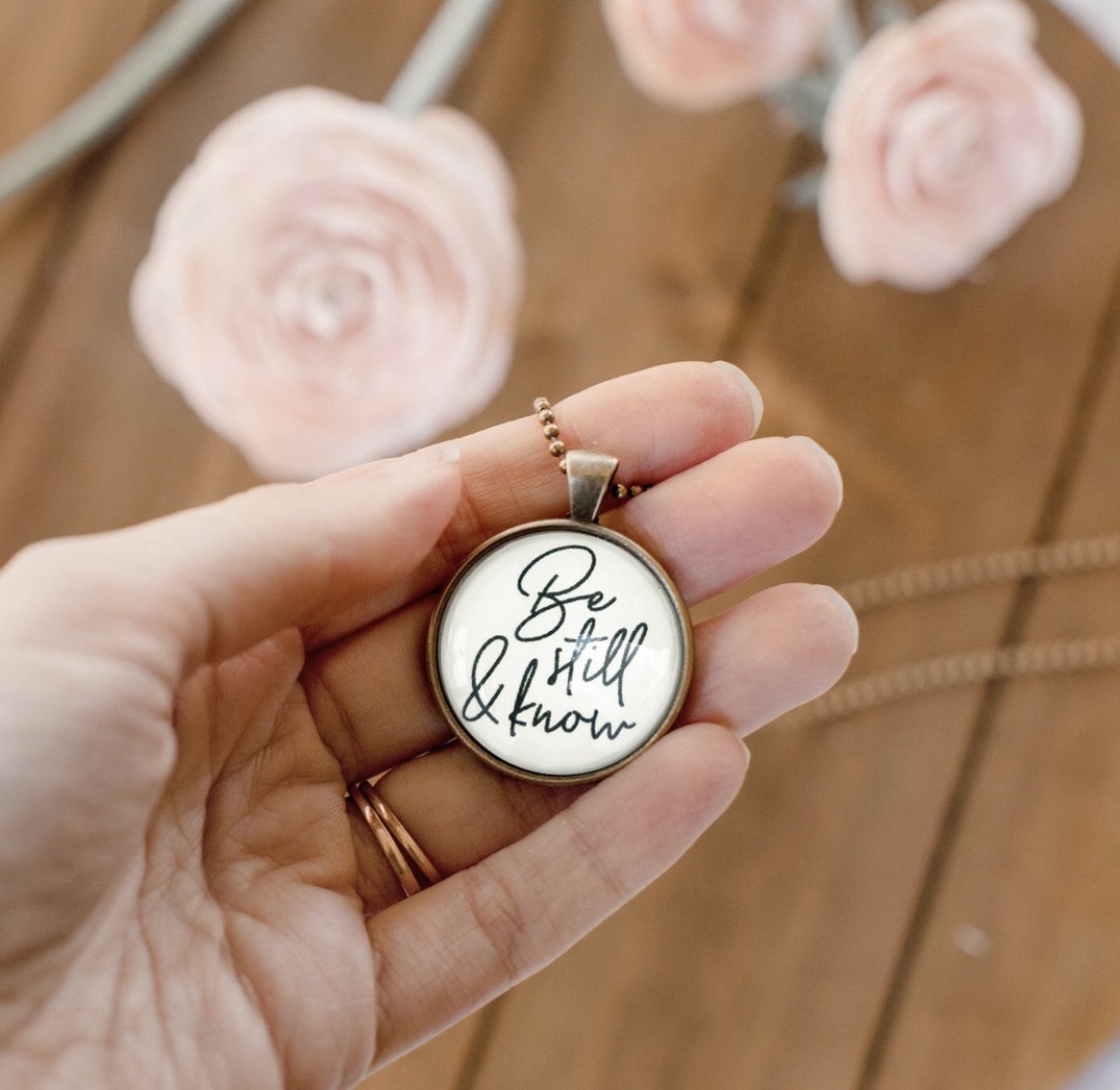 Hand holding a round pendant with handwritten text, background of pink roses and wooden surface.
