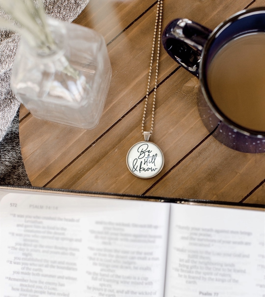 Necklace with round pendant, cup of coffee, glass jar, and open book on wooden surface.
