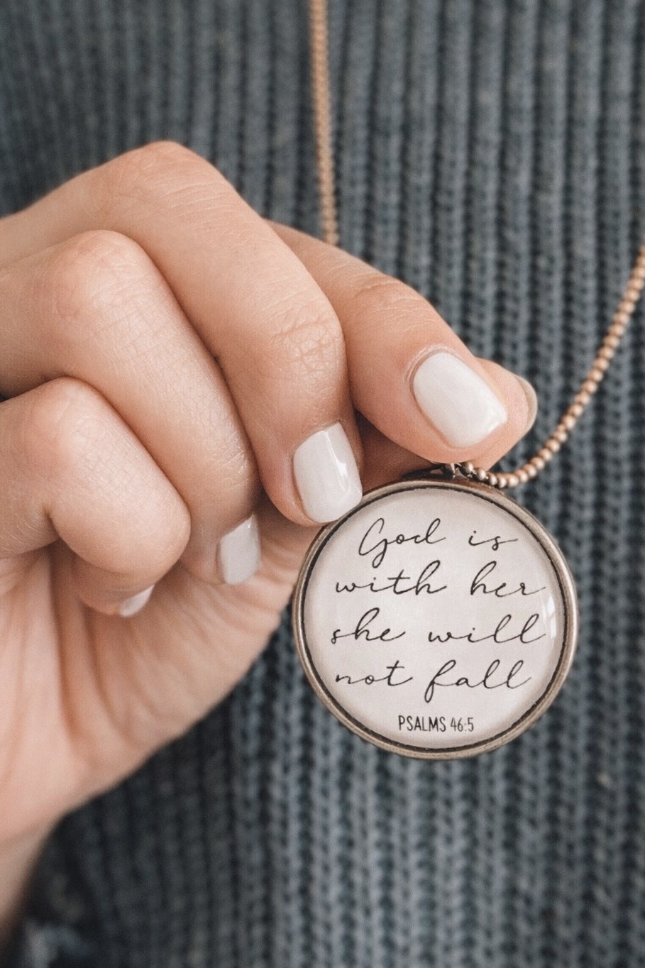 Person holding a round pendant with handwritten Bible verse, wearing a textured gray sweater, fingernails painted white.