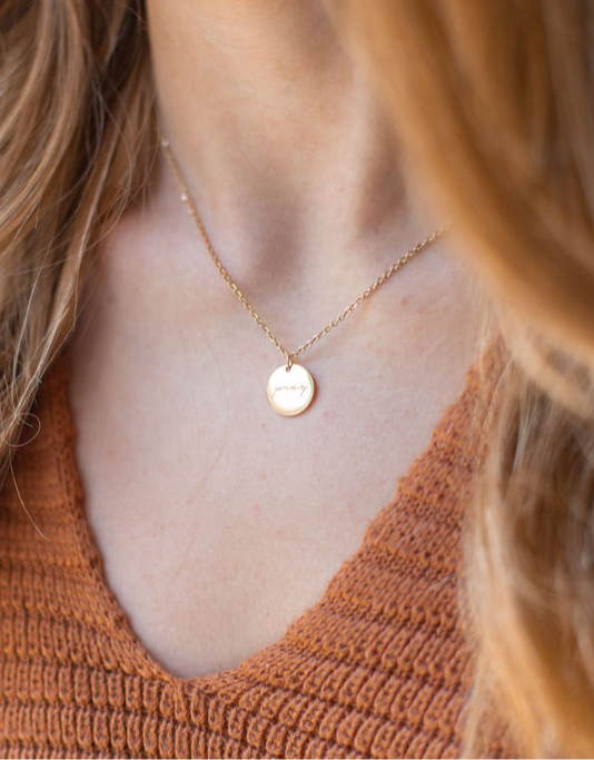 Close-up of a person's neck and chest wearing a gold necklace with a round pendant, and a rust-colored knitted top.