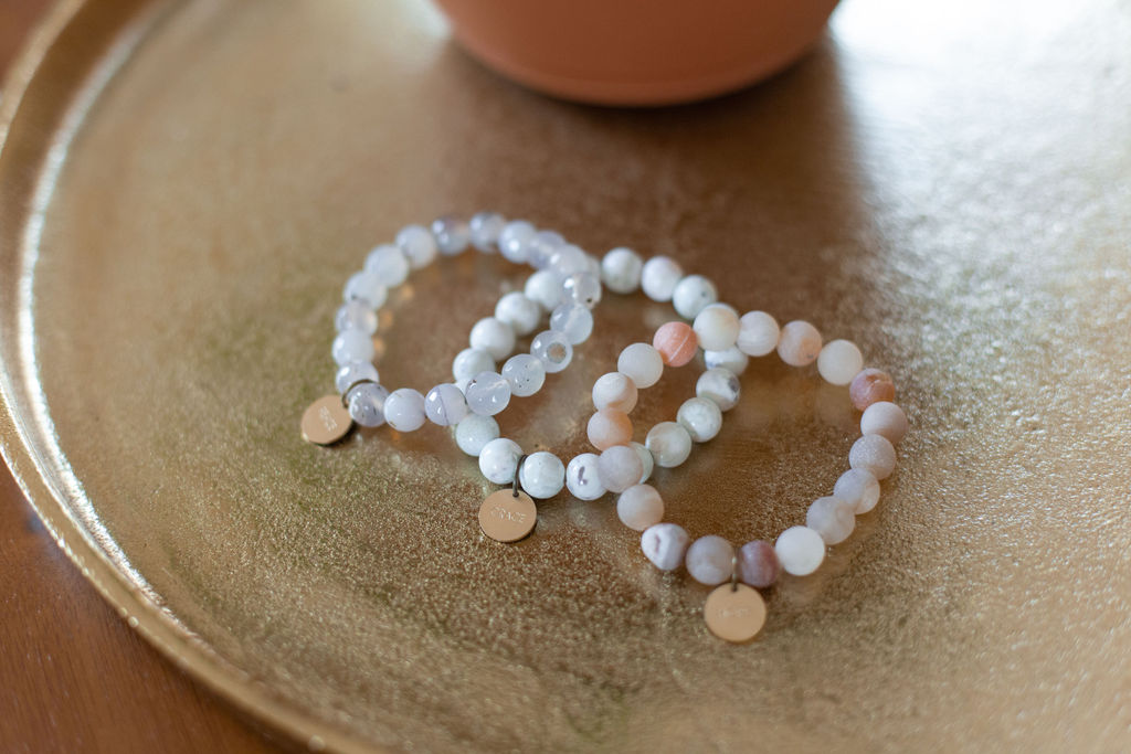 Three beaded bracelets with small round charms on a textured surface, with a blurred object in the background.