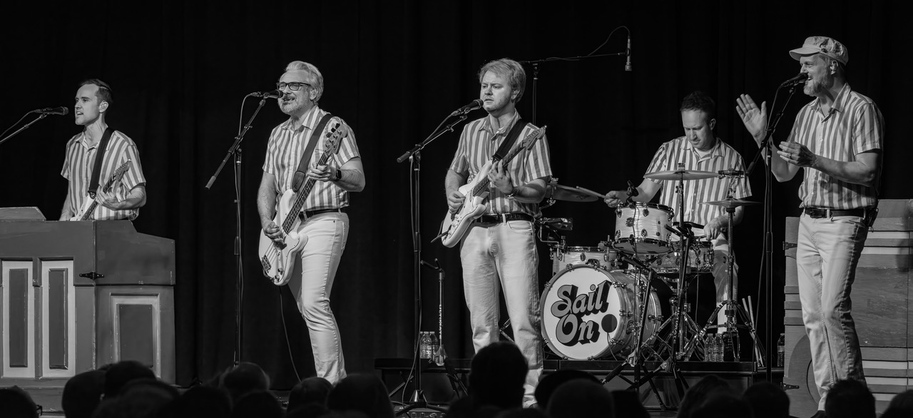 Five musicians perform on stage with instruments, wearing striped shirts, in front of a black curtain. A drum set is visible.
