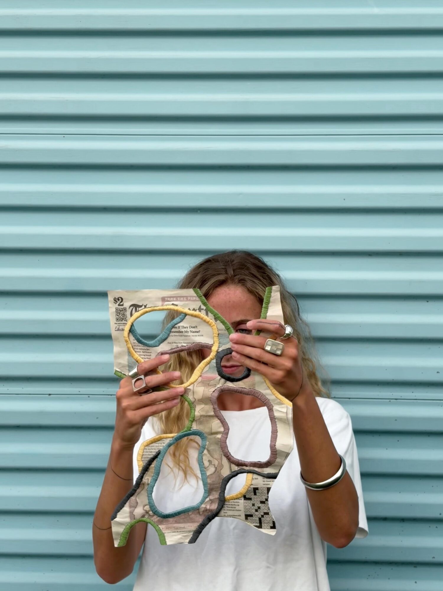 Person holding sunglasses and a patterned bag in front of a blue corrugated wall, partially covering their face.