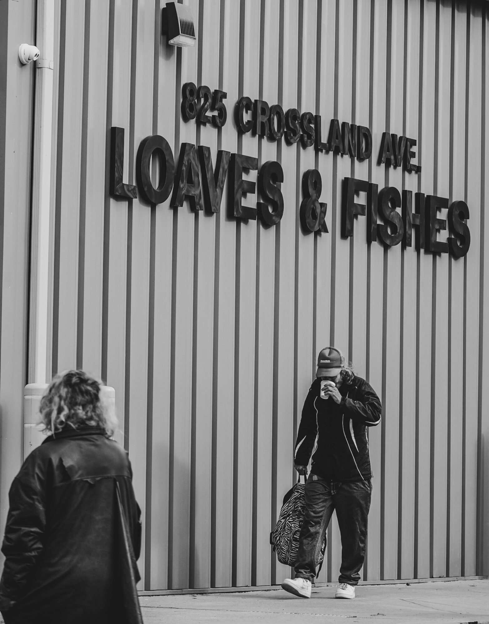 Person taking a photo outside a building with vertical metal siding and a sign reading 'LOAVES & FISHES'.