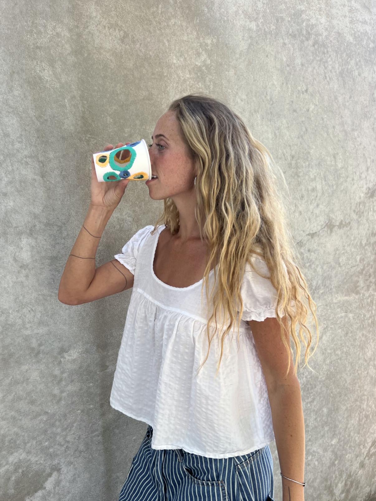 Young woman with long wavy hair drinking from a colorful mug, standing against a plain wall.