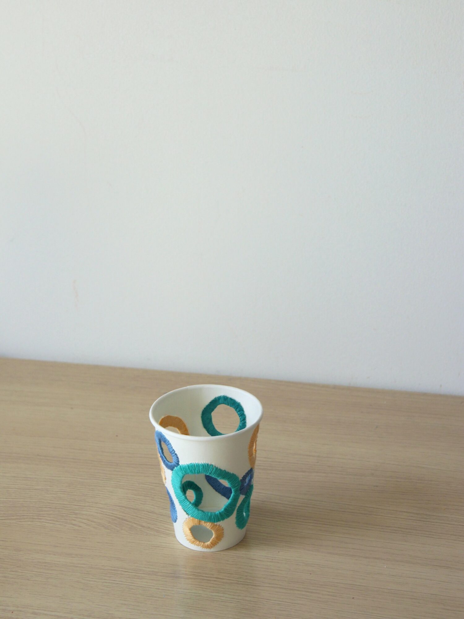 A paper cup with colorful circular patterns on a wooden surface against a plain white wall.