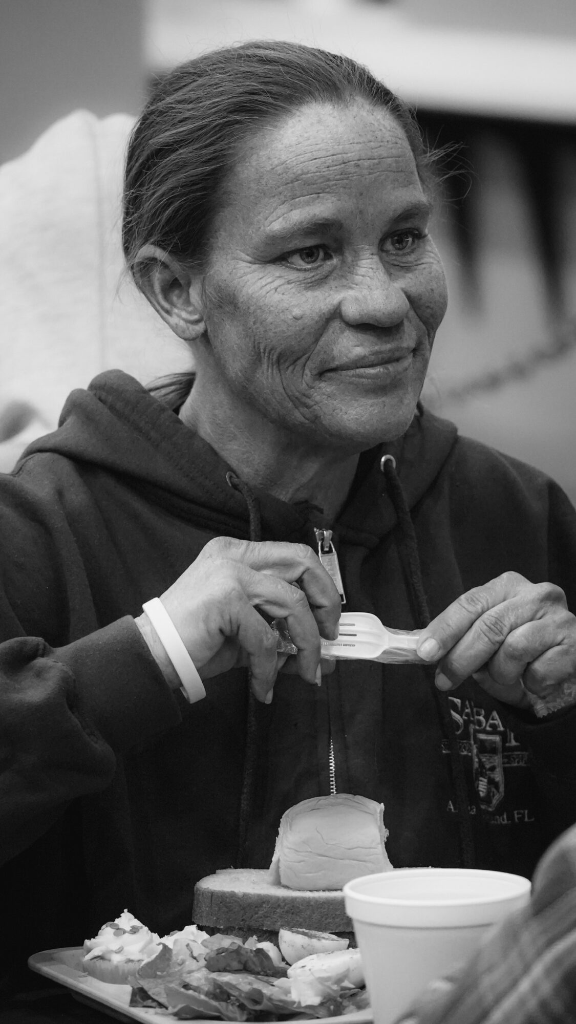 Woman with dark hair tied back, wearing a hoodie, sitting at a table with food and a cup, smiling slightly.