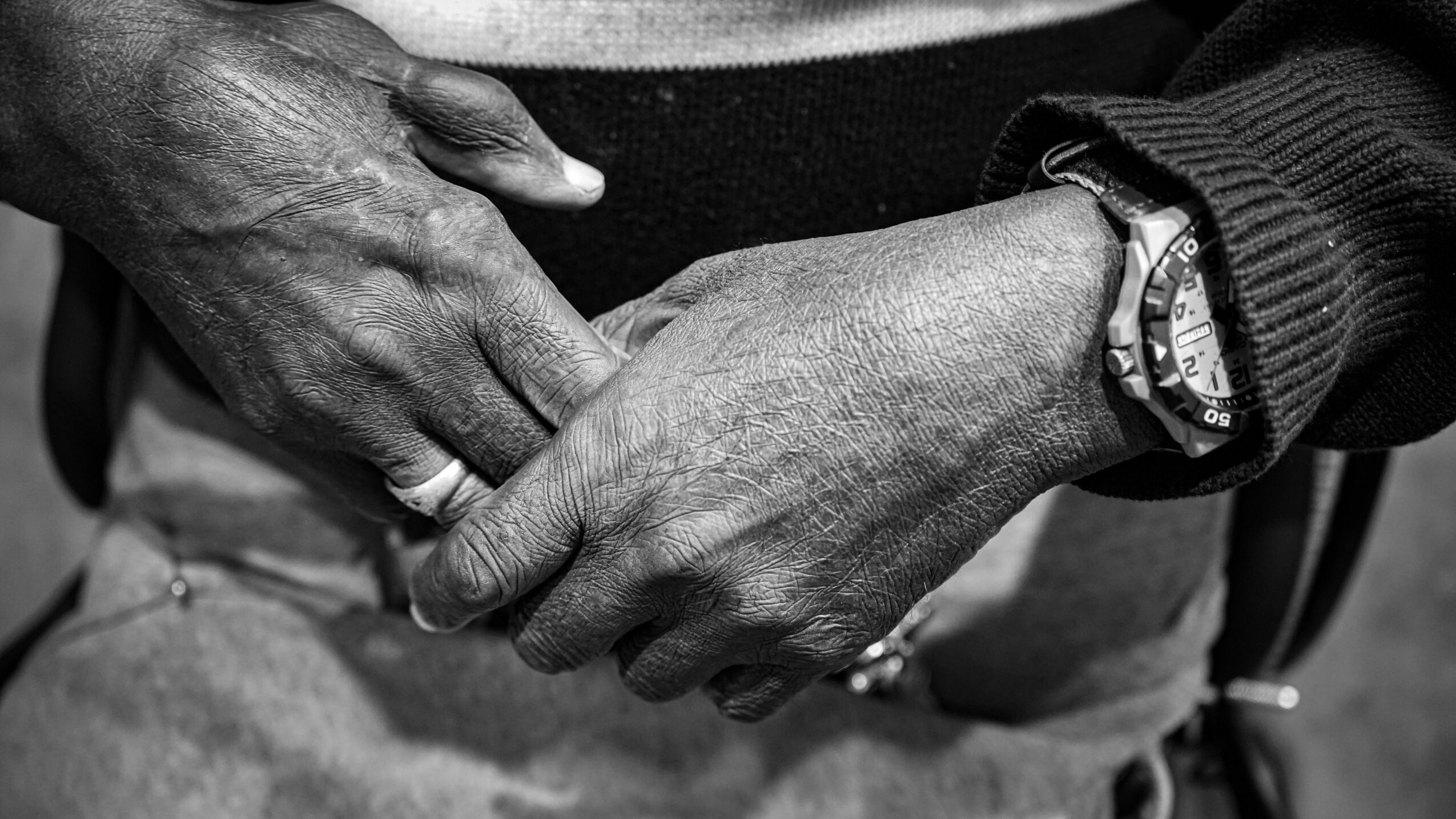 Two hands holding each other, one appears older with visible veins, the other younger, wearing a watch, in black and white.