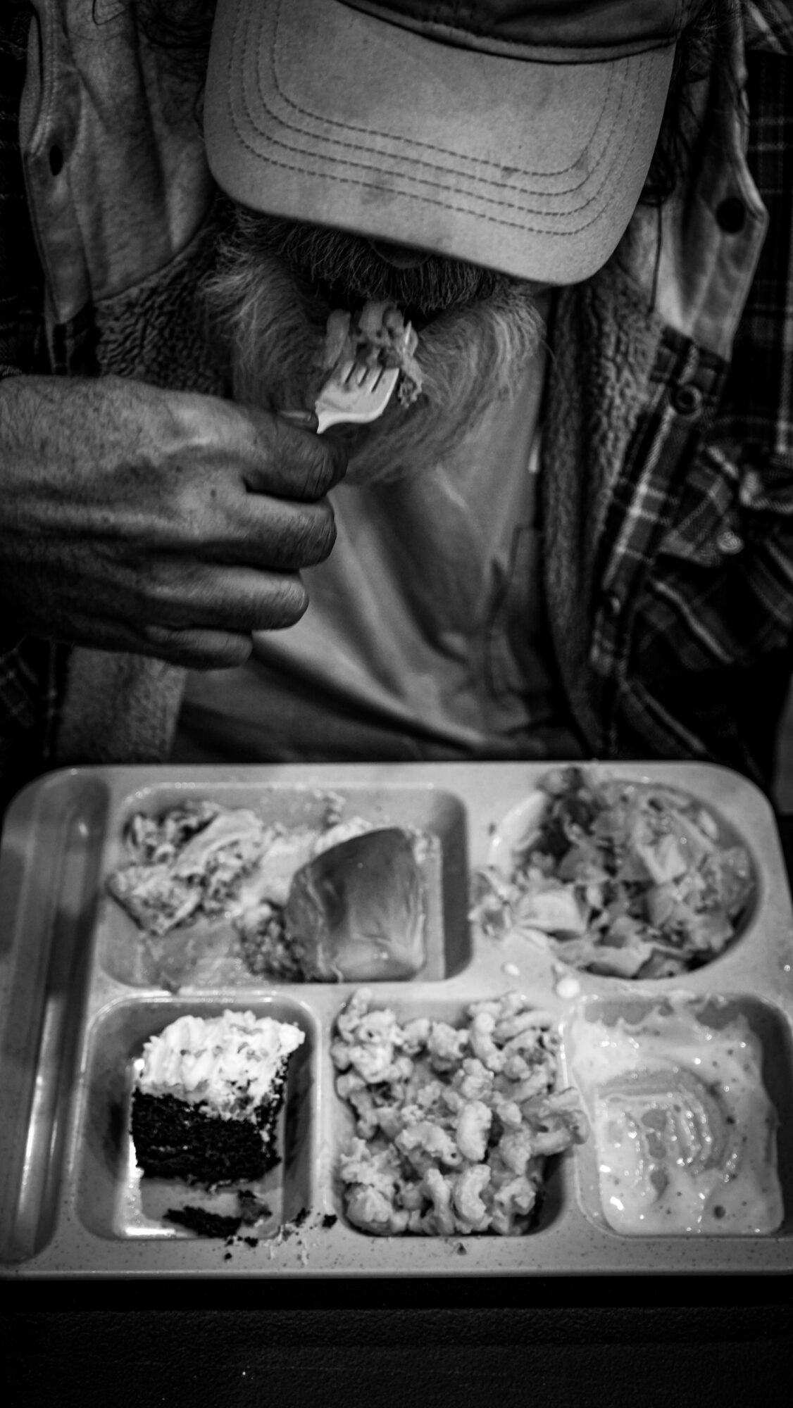 Person holding a small object, wearing a cap and layered clothing, with a tray of food in front, in black and white.