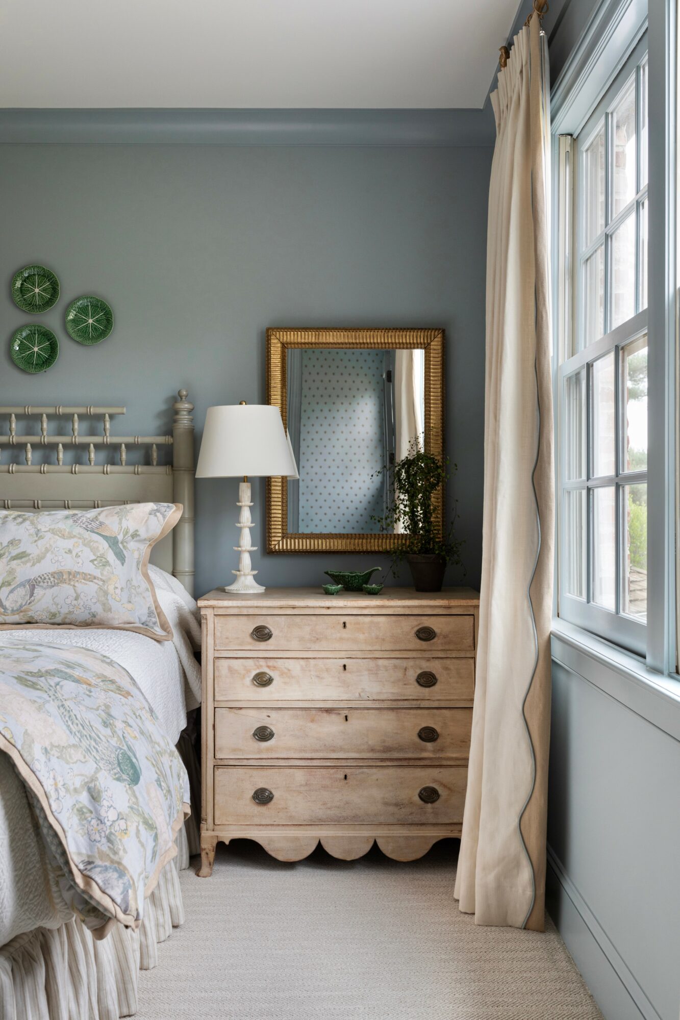 Bedroom corner with dresser, mirror, lamp, and window with curtains, light blue wall, and decorative plates.