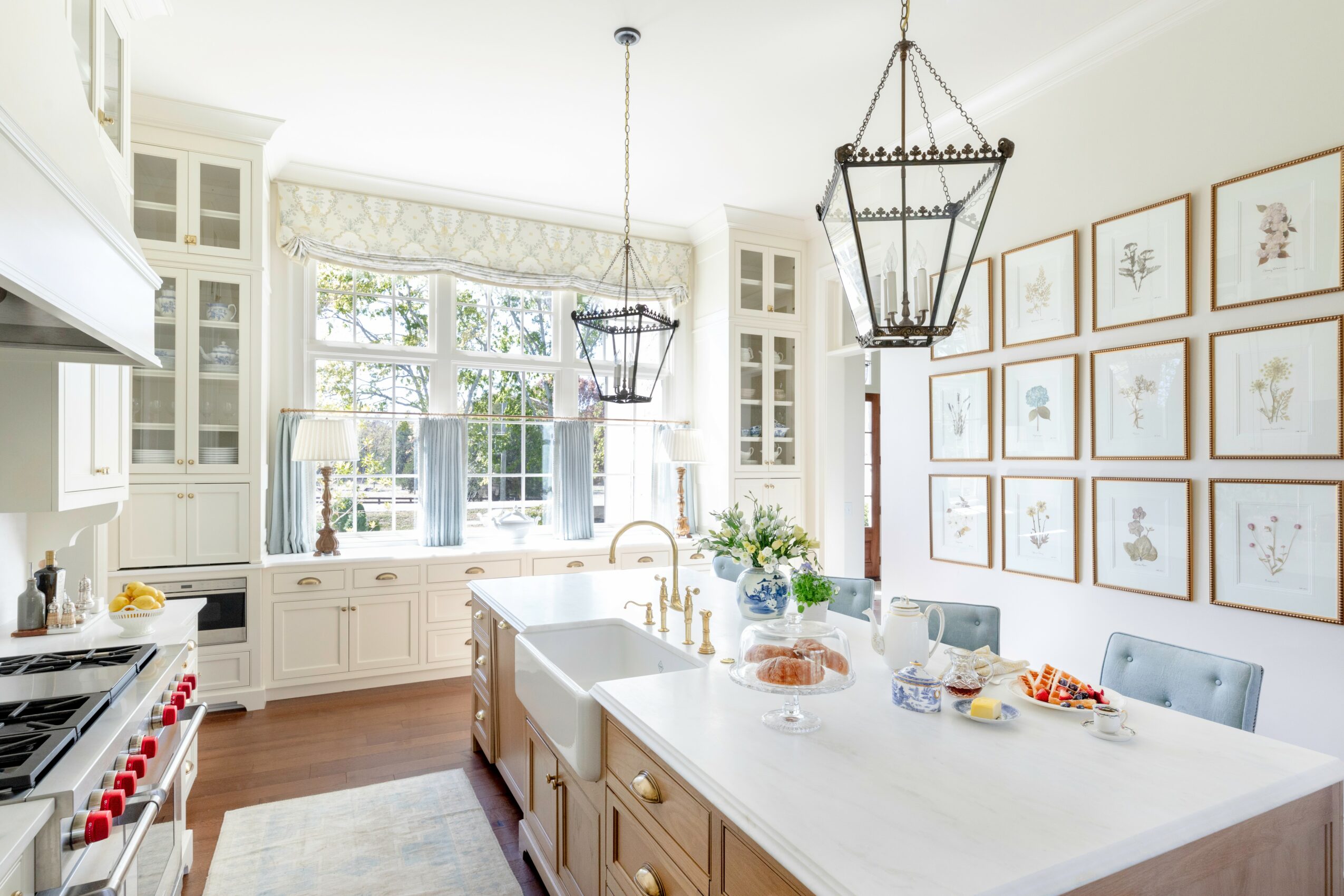 Bright kitchen with large window, white cabinets, island, and framed botanical prints on wall, decorated with flowers and food on island.
