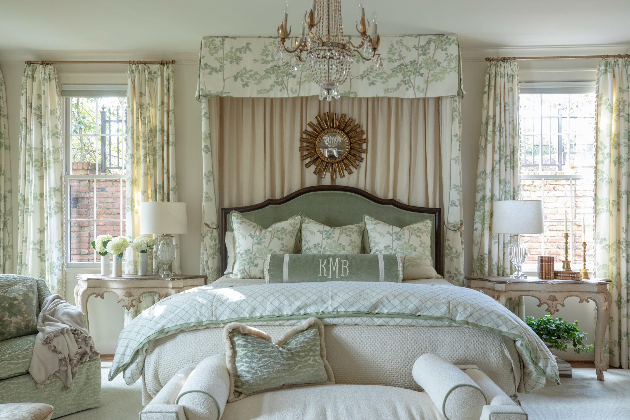 Upright bedroom with a large bed, chandelier, two windows with curtains, and bedside tables with lamps.