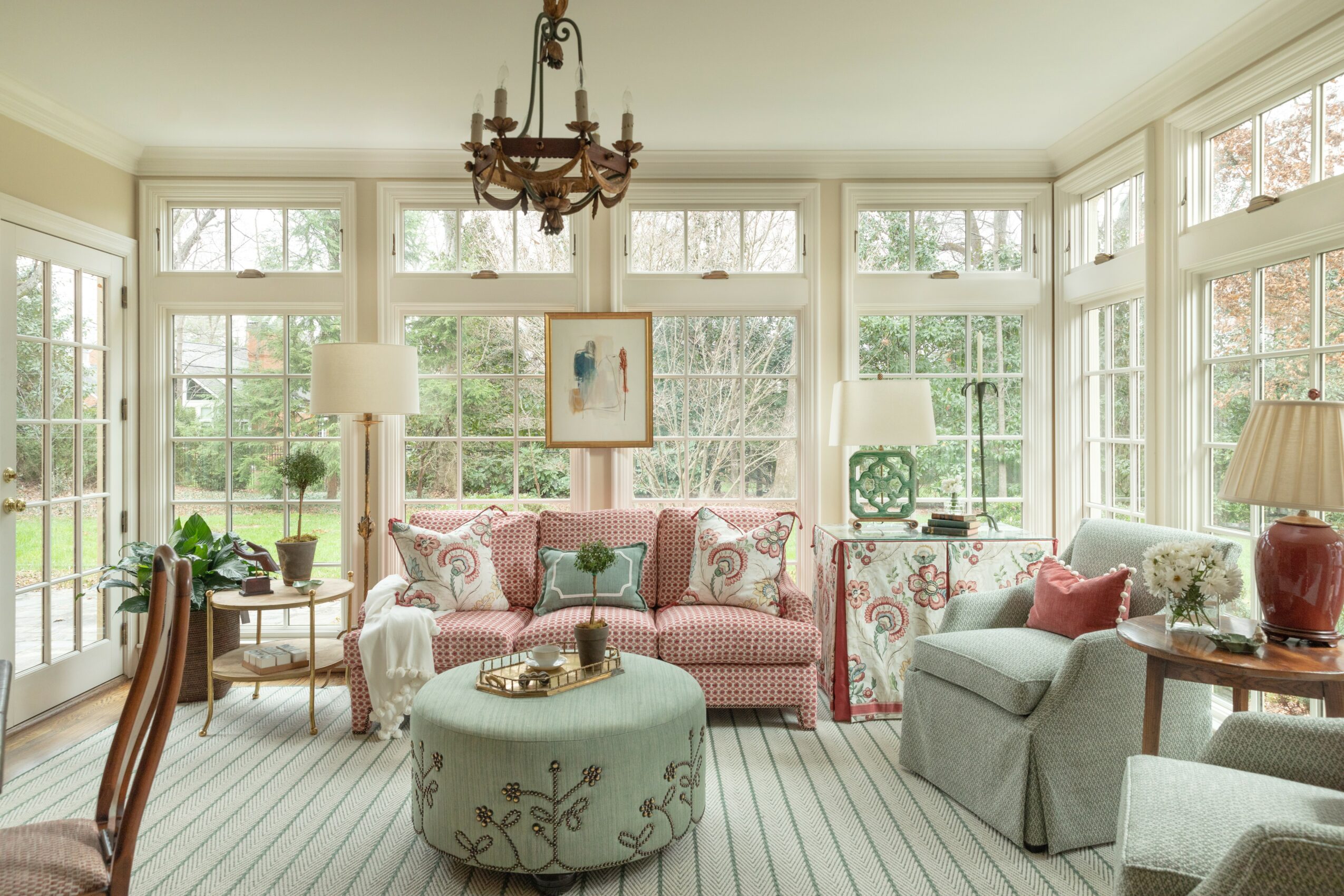 Living room with large windows, pink sofa, armchair, chandelier, and various lamps and plants, bright and airy space.