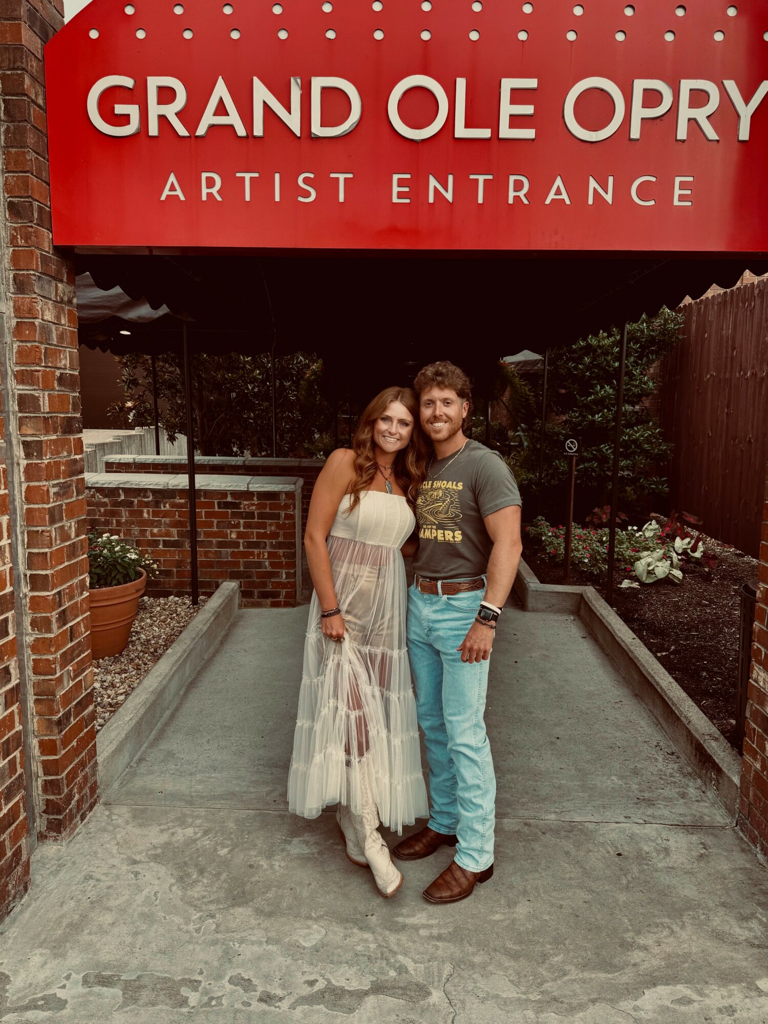 A man and woman stand together outside at the Grand Ole Opry, smiling, with a red sign above them reading 'Grand Ole Opry Artist Entrance'.